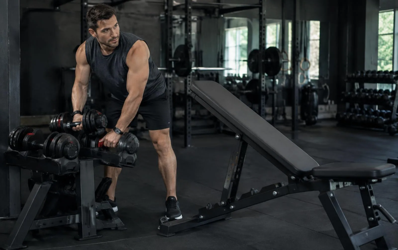A man setting adjustable dumbbells beside an incline bench.